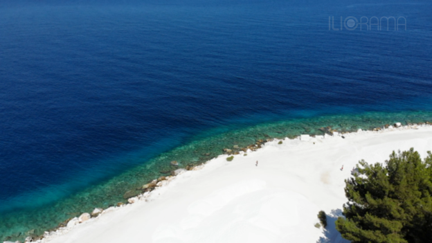 Marble beach drone shot with marble sand, blue sea, pine trees